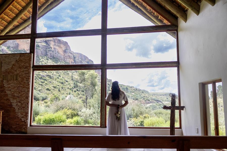 A bride standing before the chapel’s large window overlooking green slopes and sandstone mountains