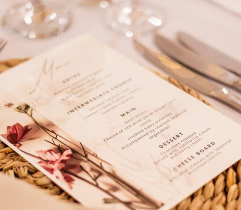 A printed wedding menu with flowers on a woven placemat beside polished cutlery