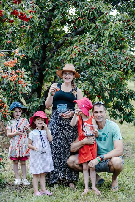 Two adults and three children holding cherries beneath fruit-laden trees in an orchard